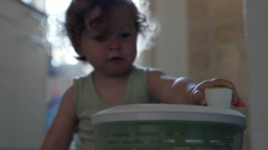 Bebek, mutfak zemininde oturmuş ev eşyalarıyla uğraşırken salata fırıldağı kapağında el sıkışıyor.