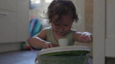 Bebek mutfak zemininde oynarken salata fırıldağı kapağını kaldırıyor, odaklanmış ifade ve eller konteynırla iç mekanda etkileşime giriyor