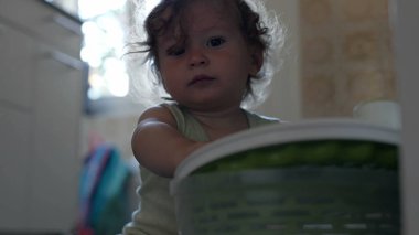 Bebek, mutfak zemininde oturmuş ev eşyalarıyla uğraşırken salata fırıldağı kapağında el sıkışıyor.