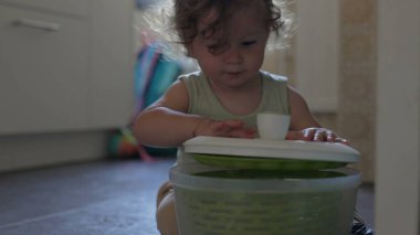 Bebek mutfak zemininde oynarken salata fırıldağı kapağını kaldırıyor, odaklanmış ifade ve eller konteynırla iç mekanda etkileşime giriyor