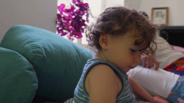 Küçük çocuk koltukta dik otururken anne uzanır ve ağabeyi yastıklarda uzanır. Huzurlu, rahat bir oturma odasında huzurlu bir aile anı.