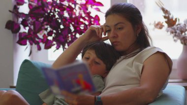 Anne kanepede oğluyla birlikte çocuk kitapları okuyor, ikisi de sayfaya odaklanmış, arka planda bitkiler ve pencereden güneş ışığı görünen rahat ev ortamları.