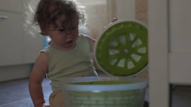 Bebek mutfak zemininde oynarken salata fırıldağı kapağını kaldırıyor, odaklanmış ifade ve eller konteynırla iç mekanda etkileşime giriyor