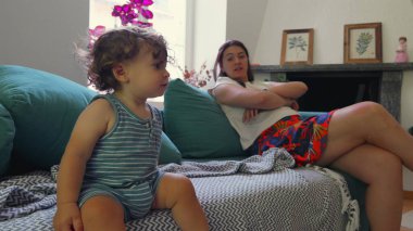 Kıvırcık saçlı çocuk oturma odasındaki kanepede dik otururken anne arkasını kolluyor. Gün ışığı ve ev dekoruyla çerçevelenmiş.