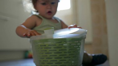 Yürüyen çocuk salata fırıldağıyla oynuyor, mutfak objesini merakla inceliyor, ellerinde büyük yeşil plastik bir kap, kapalı ev sahnesi.