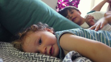 Kıvırcık saçlı bebek yastığa uzanmış, yumuşak gülüşüyle kameraya bakıyor, ağabeyi ve annesi aydınlık, rahat bir oturma odasında dinleniyor.