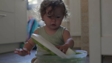 Bebek mutfak zemininde oynarken salata fırıldağı kapağını kaldırıyor, odaklanmış ifade ve eller konteynırla iç mekanda etkileşime giriyor