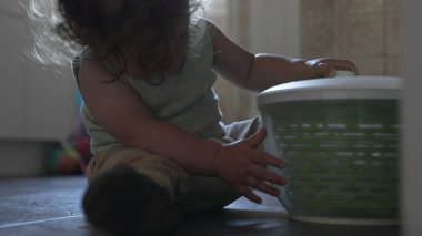 Bebek mutfakta oturmuş salata çeviricisiyle oynuyor. İki elinde yeşil plastik kapları inceliyor.