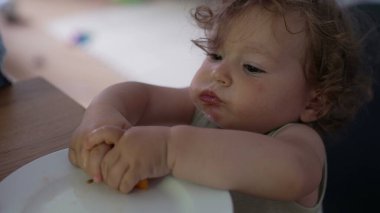 Bebek elinde kavun parçaları tutarken ve masada otururken büyük yanaklarla çiğnerken meraklı ve odaklanmış yemek anlarını kapatıyor.