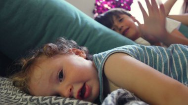 Kıvırcık saçlı bebek yastığa uzanmış, yumuşak gülüşüyle kameraya bakıyor, ağabeyi ve annesi aydınlık, rahat bir oturma odasında dinleniyor.