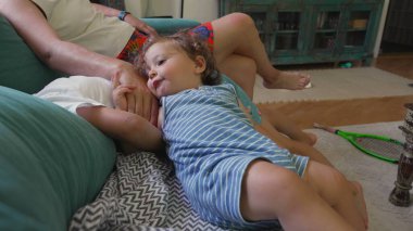 Kıvırcık saçlı çocuk, kardeşlerinin vücudunda sırtüstü uzanmış yatarken, anne de yakınlardan, rahat ve aile varlığıyla dolu sıradan bir oturma odasından izliyor.