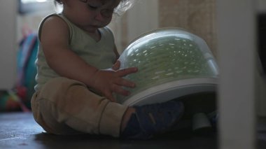 Bebek mutfakta oturmuş salata çeviricisiyle oynuyor. İki elinde yeşil plastik kapları inceliyor.