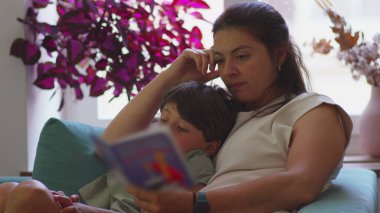 Anne kanepede oğluyla birlikte çocuk kitapları okuyor, ikisi de sayfaya odaklanmış, arka planda bitkiler ve pencereden güneş ışığı görünen rahat ev ortamları.