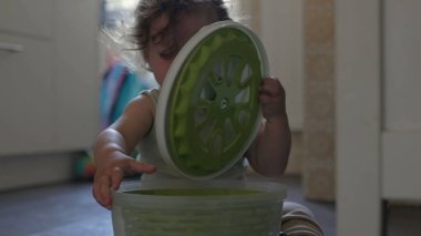 Bebek mutfak zemininde oynarken salata fırıldağı kapağını kaldırıyor, odaklanmış ifade ve eller konteynırla iç mekanda etkileşime giriyor