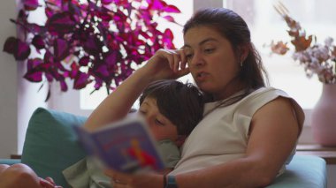 Anne kanepede oğluyla birlikte çocuk kitapları okuyor, ikisi de sayfaya odaklanmış, arka planda bitkiler ve pencereden güneş ışığı görünen rahat ev ortamları.