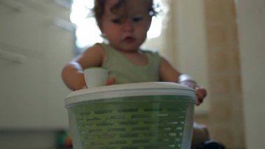 Yürüyen çocuk salata fırıldağıyla oynuyor, mutfak objesini merakla inceliyor, ellerinde büyük yeşil plastik bir kap, kapalı ev sahnesi.