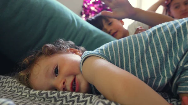 Kıvırcık saçlı bebek yastığa uzanmış, yumuşak gülüşüyle kameraya bakıyor, ağabeyi ve annesi aydınlık, rahat bir oturma odasında dinleniyor.