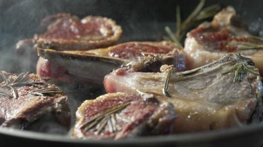 Kuzu pirzolası tavada pişiyor, biberiye ve hafif dumanla, yüzeyde altın kabuk gelişiyor, yağ ve kavrulmuş kenarlar parlıyor.