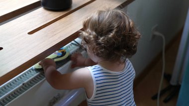 Toddler masanın altındaki elektrik prizi alanını araştırıyor, ev eşyaları ve kablo ile çerçevelenmiş şekilde yerde dururken sokete doğru uzanıyor.