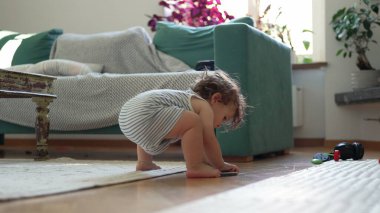 Küçük çocuk oturma odasında yerde oturuyor, tek koluyla dengede duruyor, bacakları kaldırılmış, rahat mobilyalarla çevrili, kanepe, halı ve dağınık oyuncaklar da dahil, iç mekan ışıklarında.