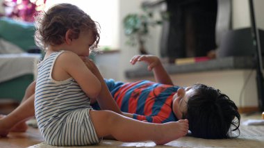 Bebek ve ağabeyi oturma odasında yerde oynuyor, oyuncaklar ve paspaslarla dinlenme anında gülüyor ve etkileşime geçiyorlar.