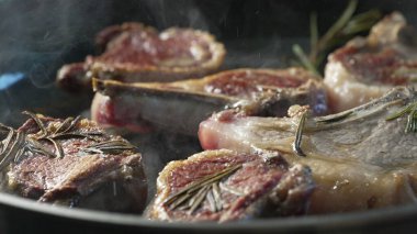 Kuzu pirzolası tavada pişiyor, biberiye ve hafif dumanla, yüzeyde altın kabuk gelişiyor, yağ ve kavrulmuş kenarlar parlıyor.