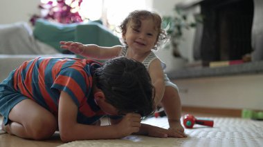Oturma odasındaki küçük çocukla büyük kardeş arasında neşeli bir etkileşim, yerde sevgi dolu bir kucaklaşma yaşarlarken samimi bir bağ ve denge anı.