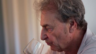Yaşlı adam kadehten son yudum beyaz şarap içer, dudakları kenardan büzüşür kapalı mekan manzarası sırasında yakın profil görüntüsünde.