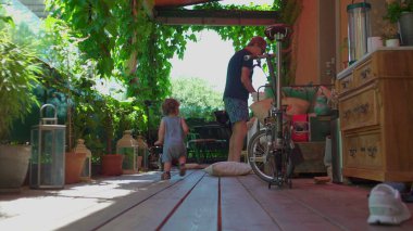 Kıvırcık saçlı bir çocuk gölgeli terasta yürürken etrafı bitkilerle, mobilyalarla ve katlanır bisikletle çevrili.