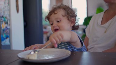 Kıvırcık saçlı bebek yemek yedikten sonra yemek masasında kaşık ve tabakla oynuyor, merak ediyor ve aile mutfağında gün ışığının yumuşak olduğu bir ortamda odaklanıyor.