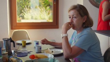 Yaşlı bir kadın kahvaltı masasında muz yerken kırmızı elbiseli genç bir kadın yakınlarda duruyor, sabah ışığı rahat mutfak düzenini dolduruyor.