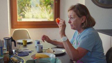 Yaşlı bir kadın kahvaltı masasında muz yerken kırmızı elbiseli genç bir kadın yakınlarda duruyor, sabah ışığı rahat mutfak düzenini dolduruyor.