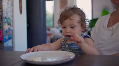 Kıvırcık saçlı bebek yemek yedikten sonra yemek masasında kaşık ve tabakla oynuyor, merak ediyor ve aile mutfağında gün ışığının yumuşak olduğu bir ortamda odaklanıyor.