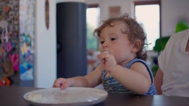 Kıvırcık saçlı bebek yemek yedikten sonra yemek masasında kaşık ve tabakla oynuyor, merak ediyor ve aile mutfağında gün ışığının yumuşak olduğu bir ortamda odaklanıyor.