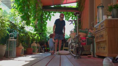 Kıvırcık saçlı bir çocuk gölgeli terasta yürürken etrafı bitkilerle, mobilyalarla ve katlanır bisikletle çevrili.