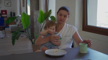 Anne kucağında bebekle sabah kahvaltısı yaparken kucağında bebek kaşığıyla büyük pencerenin yanındaki aile sofrasında oynarken bardaktan yudumluyor.