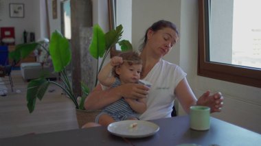 Anne kucağında bebekle sabah kahvaltısı yaparken kucağında bebek kaşığıyla büyük pencerenin yanındaki aile sofrasında oynarken bardaktan yudumluyor.