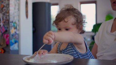Kıvırcık saçlı bebek yemek yedikten sonra yemek masasında kaşık ve tabakla oynuyor, merak ediyor ve aile mutfağında gün ışığının yumuşak olduğu bir ortamda odaklanıyor.