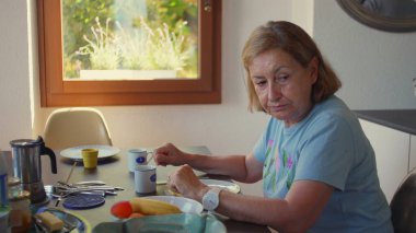 Yaşlı bir kadın kahvaltı masasında muz yerken kırmızı elbiseli genç bir kadın yakınlarda duruyor, sabah ışığı rahat mutfak düzenini dolduruyor.