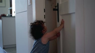 Bebek çabayla kapı tokmağına uzanıyor, modern evdeki kapıyı açmaya çalışıyor, parmak uçlarında gelişigüzel duruyor.