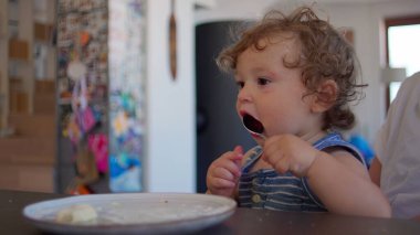 Kıvırcık saçlı bebek, geniş ağızlı kaşığı hevesle yalıyor, sıcak ev mutfağında yemek saatinde aile yemek masasında oturuyor.