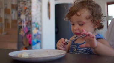 Kıvırcık saçlı bebek, geniş ağızlı kaşığı hevesle yalıyor, sıcak ev mutfağında yemek saatinde aile yemek masasında oturuyor.