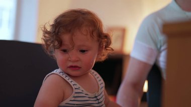 Yeni yürümeye başlayan çocuk, kollarını masaya uzatmış, gözleri yaşlı ve ağzı açık, sıkıntılı bir ifadeyle, sıcak ev içinde kıvırcık saç çerçevesi profili çiziyor.