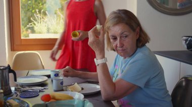 Yaşlı bir kadın kahvaltı masasında muz yerken kırmızı elbiseli genç bir kadın yakınlarda duruyor, sabah ışığı rahat mutfak düzenini dolduruyor.