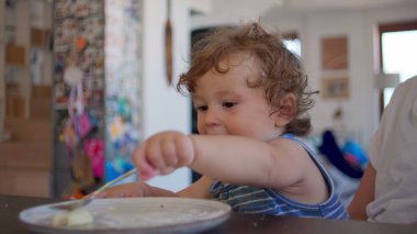 Kıvırcık saçlı bebek, geniş ağızlı kaşığı hevesle yalıyor, sıcak ev mutfağında yemek saatinde aile yemek masasında oturuyor.