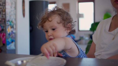 Kıvırcık saçlı bebek yemek yedikten sonra yemek masasında kaşık ve tabakla oynuyor, merak ediyor ve aile mutfağında gün ışığının yumuşak olduğu bir ortamda odaklanıyor.