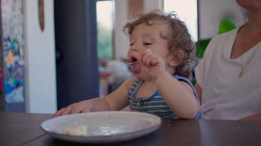 Kıvırcık saçlı bebek yemek yedikten sonra yemek masasında kaşık ve tabakla oynuyor, merak ediyor ve aile mutfağında gün ışığının yumuşak olduğu bir ortamda odaklanıyor.