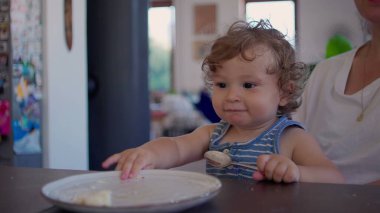 Kıvırcık saçlı bebek yemek yedikten sonra yemek masasında kaşık ve tabakla oynuyor, merak ediyor ve aile mutfağında gün ışığının yumuşak olduğu bir ortamda odaklanıyor.