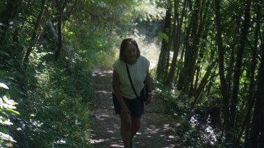 Yaşlı kadın orman yolunda yürüyor, destek için el ele yokuş yukarı yürüyor, yaz boyunca ağaçlar ve yeşil bitkilerle çevrili.
