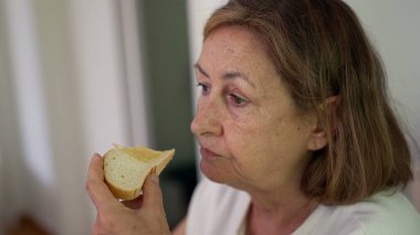 Beyaz duvar arkasında otururken sandviçi ısıran, tarafsız tişört giyen ve yemeğine odaklanmış yaşlı bir kadının yakın çekimi.